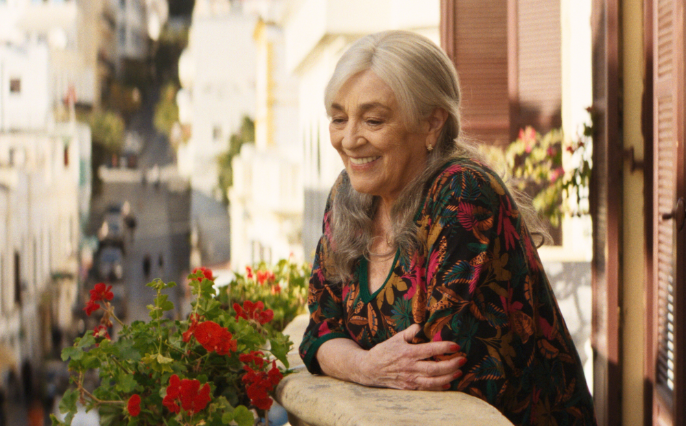 A smiling older woman leans on a balcony railing with red geraniums, in a still from the film Calle Málaga
